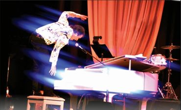 Performer balancing on a white grand piano on stage.