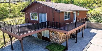 A brown house elevated on stilts with a spacious wraparound deck and railing.