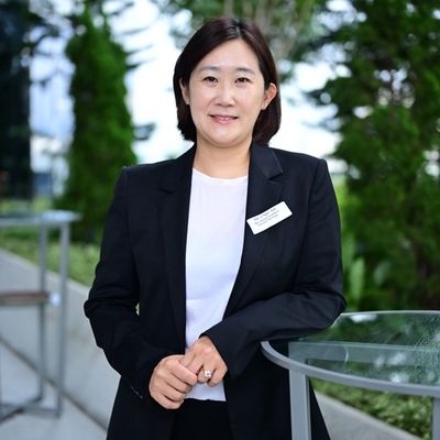 A professional woman in a black blazer smiling outdoors near greenery.
