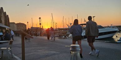 Couple walking by a marina during sunset with boats and people around.