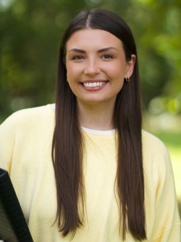 Young woman with long brown hair smiling outdoors, wearing a yellow sweater.