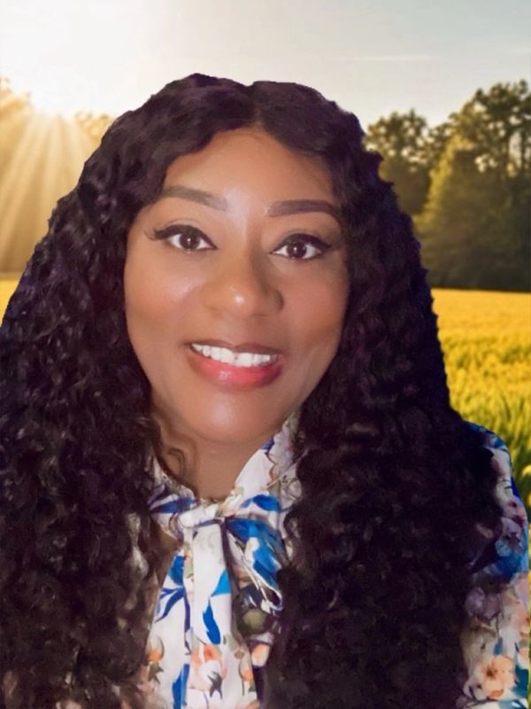 Smiling woman with long curly hair in a floral blouse, standing outdoors with sunlight and trees in the background.