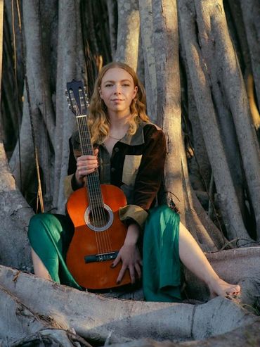 A woman with a guitar sits barefoot among large tree roots in natural light.