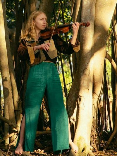 A woman plays violin barefoot in a sunlit forest.