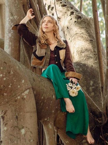 Woman in colorful outfit sitting barefoot on a large tree branch, holding flowers.