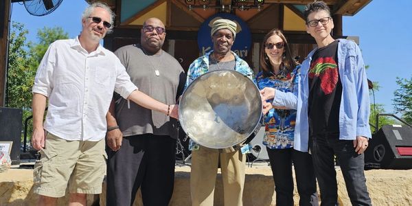 Five people on an outdoor stage holding a steel drum under a wooden canopy.