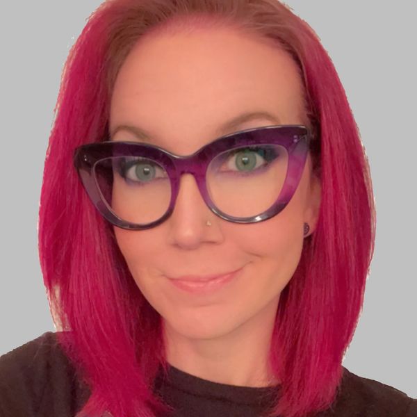 Woman wearing glasses and has pink hair smiling.