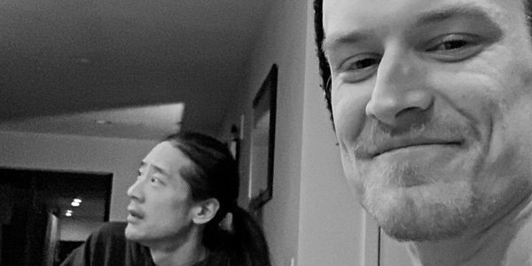 Two men sitting indoors, one smiling at the camera, the other looking away.