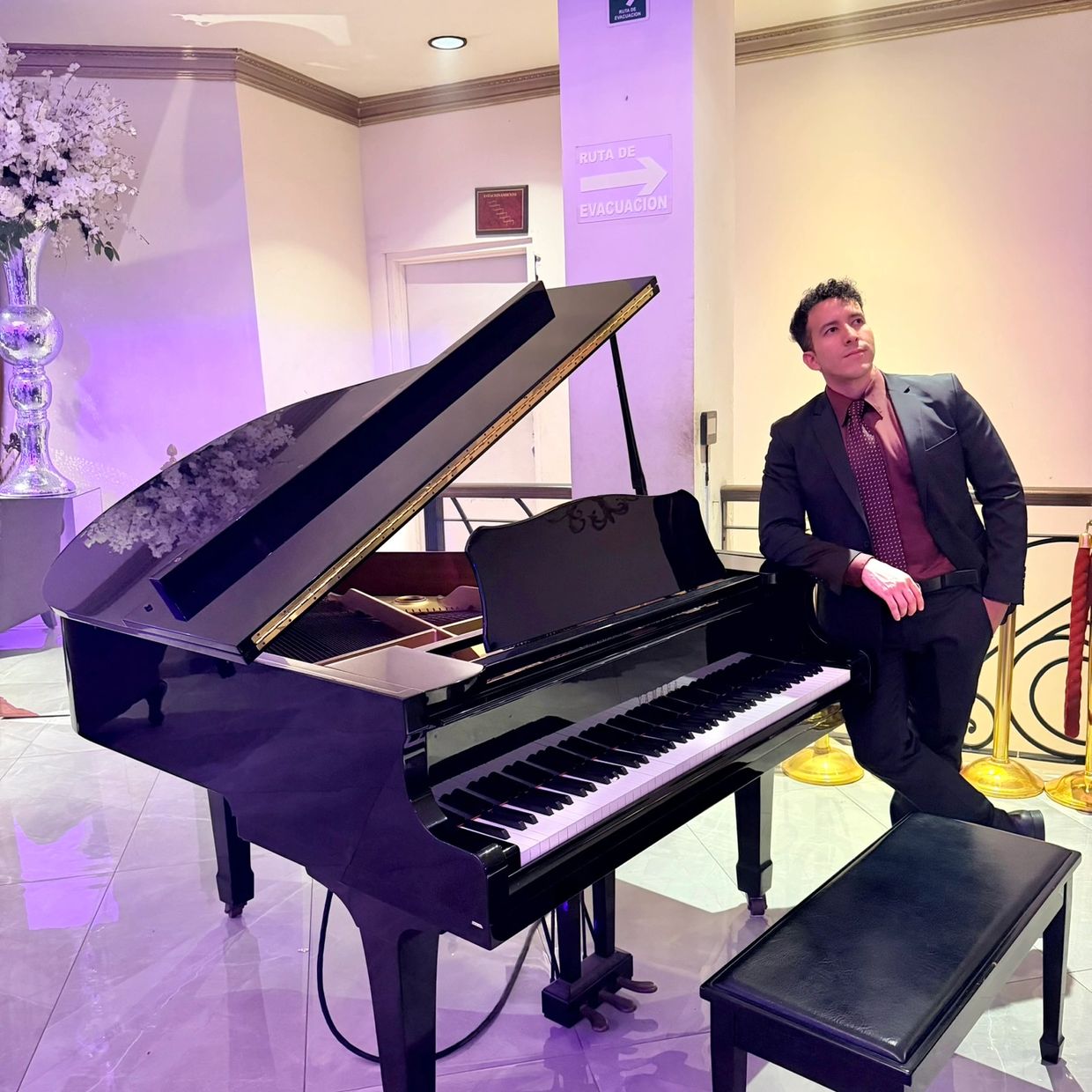 Man in formal attire leaning on a grand piano in a stylish room.