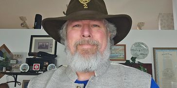 Man with gray beard wearing a hat and vest, smiling indoors.