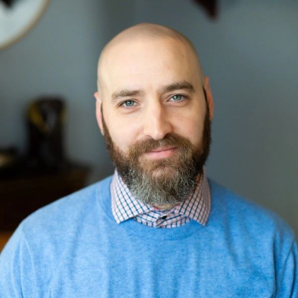 Smiling bald man with a beard wearing a blue sweater and checkered shirt.