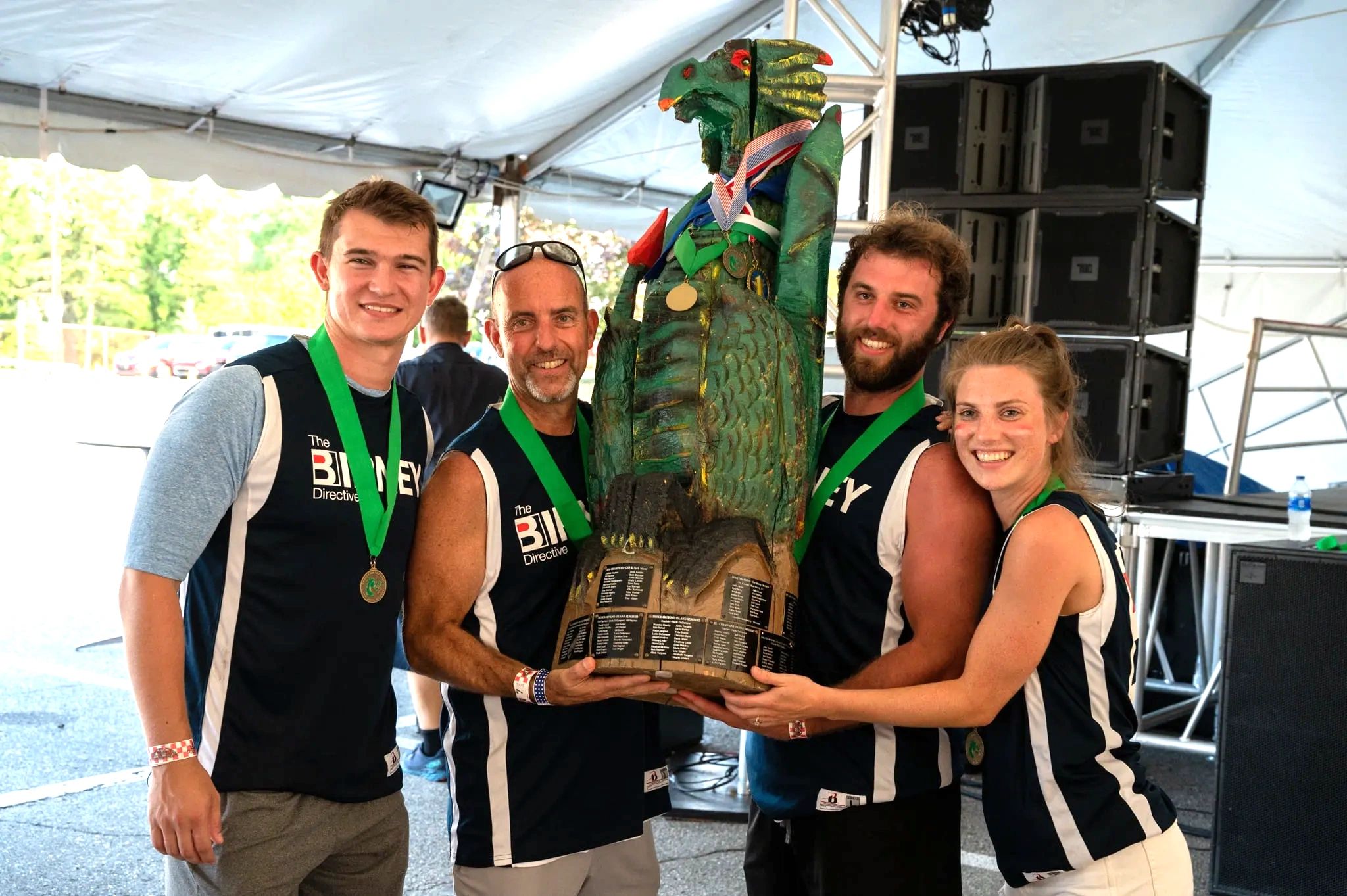Four people proudly holding a large green trophy bird under a tent.