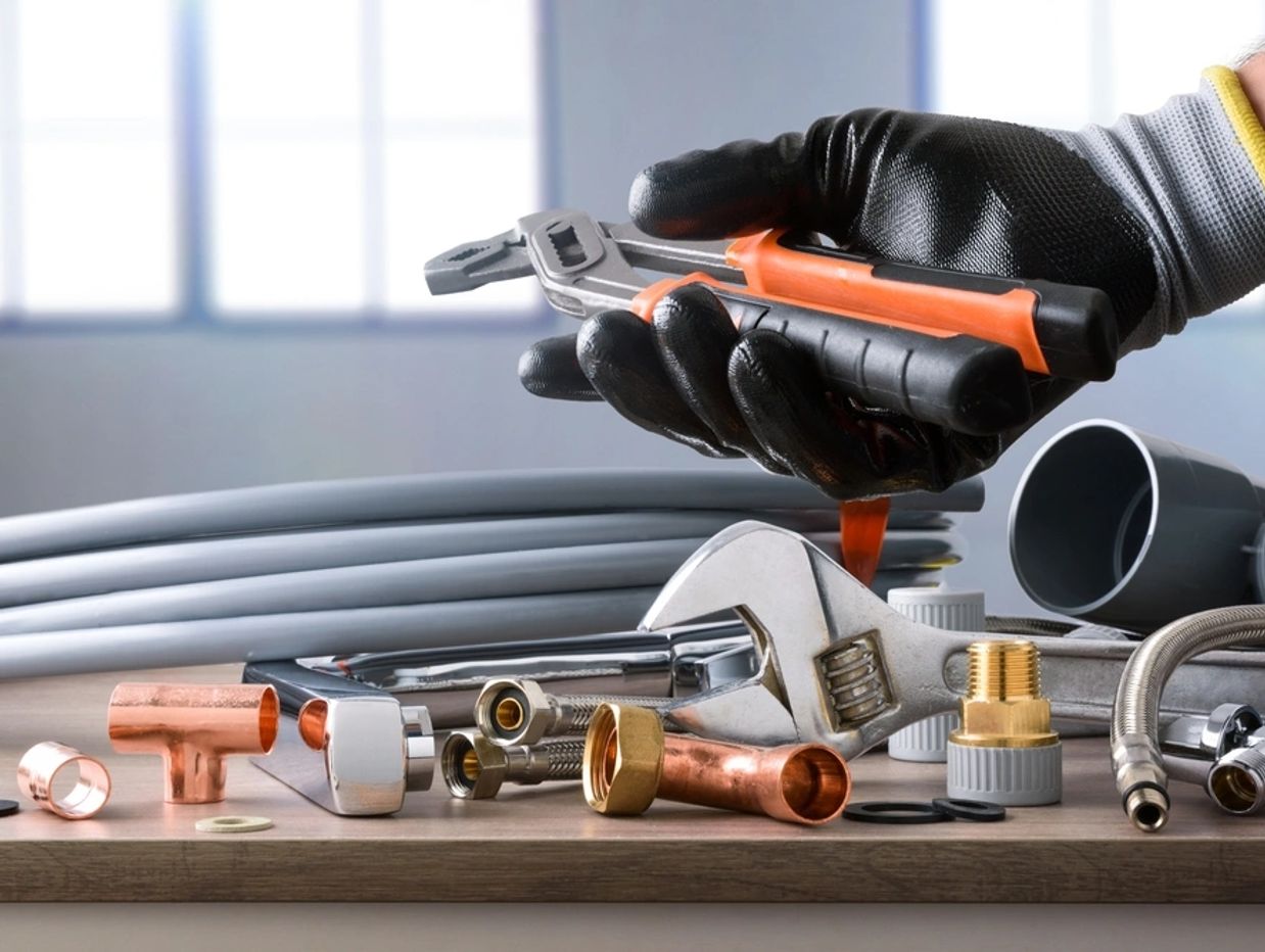 Sample of plumbing materials and tools on workbench and hand showing parrot beak pliers.