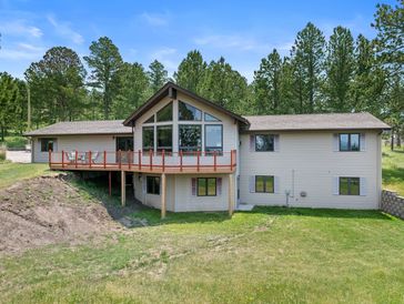 Large house with expansive deck surrounded by pine trees under a bright blue sky.