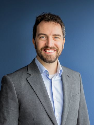 Smiling man in gray suit and blue shirt against a blue background.