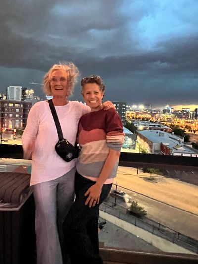 Two women smiling on a rooftop with a cityscape at dusk behind them.