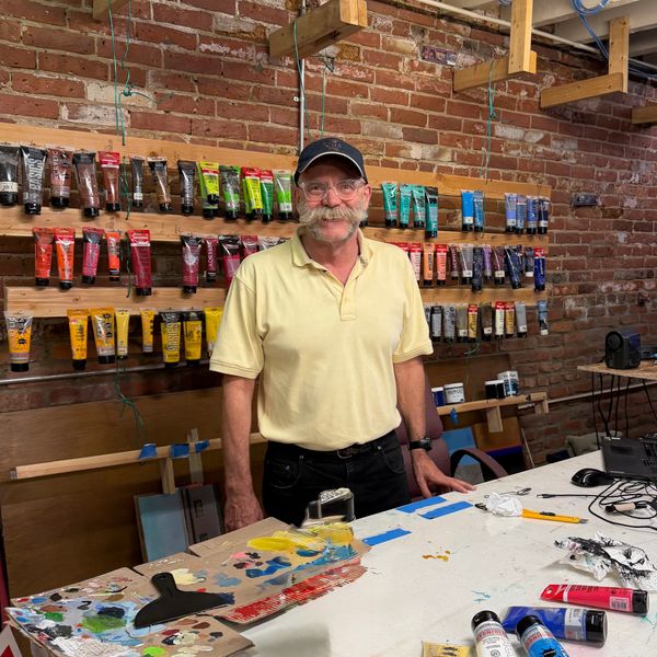 Artist in a studio surrounded by colorful paint tubes and art supplies.
