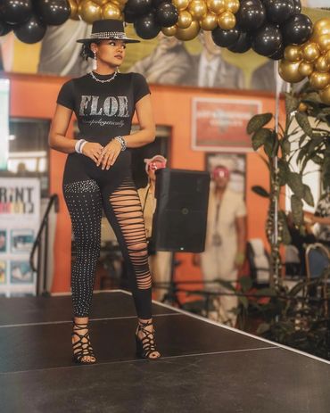 Woman in stylish black outfit and hat stands confidently on stage under black and gold balloons.