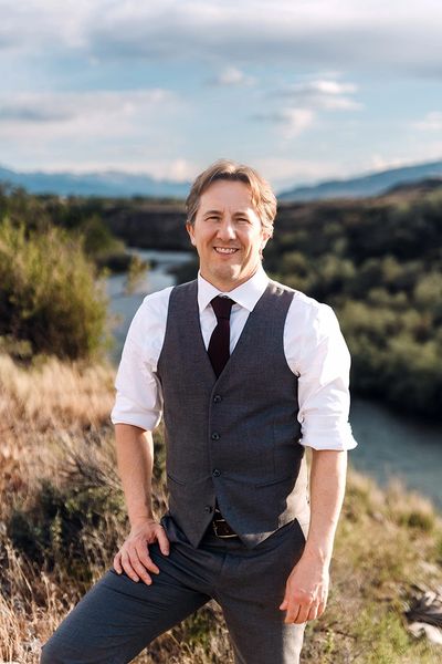 Man in formal attire posing outdoors by a river with a scenic background.