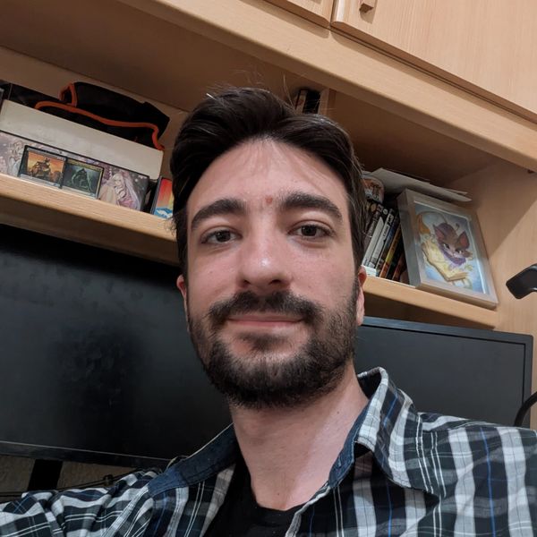 A young man with a beard smiling in front of a desk and shelves.