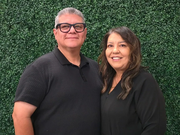 Smiling couple standing in front of a green leafy backdrop.