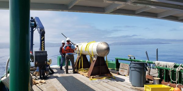 Two workers on a boat handling a large cylindrical device near the ocean.