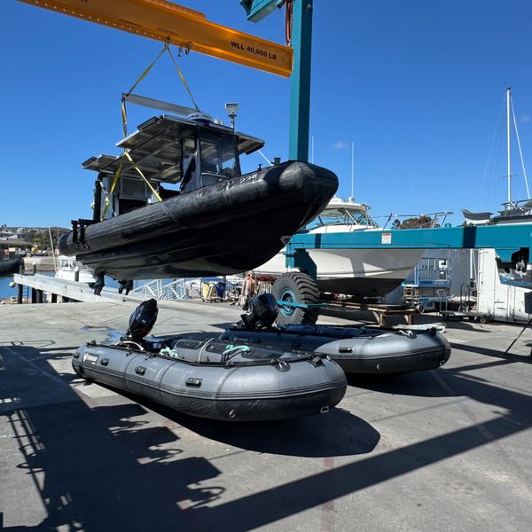 A black boat is being lifted by a crane near two inflatable boats on a sunny dock.