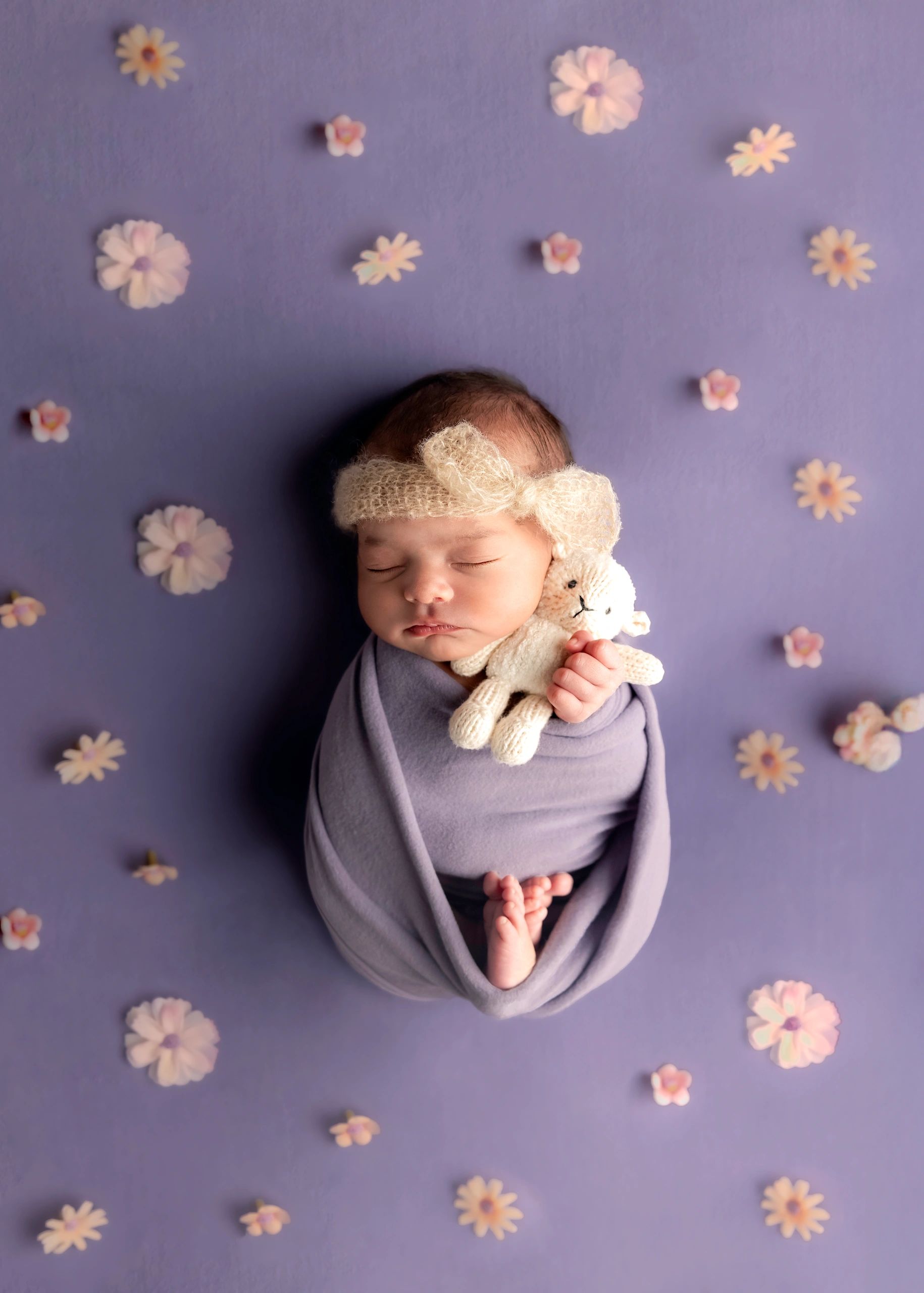 Sleeping baby wrapped in purple with a knitted headband and teddy bear on a floral background.