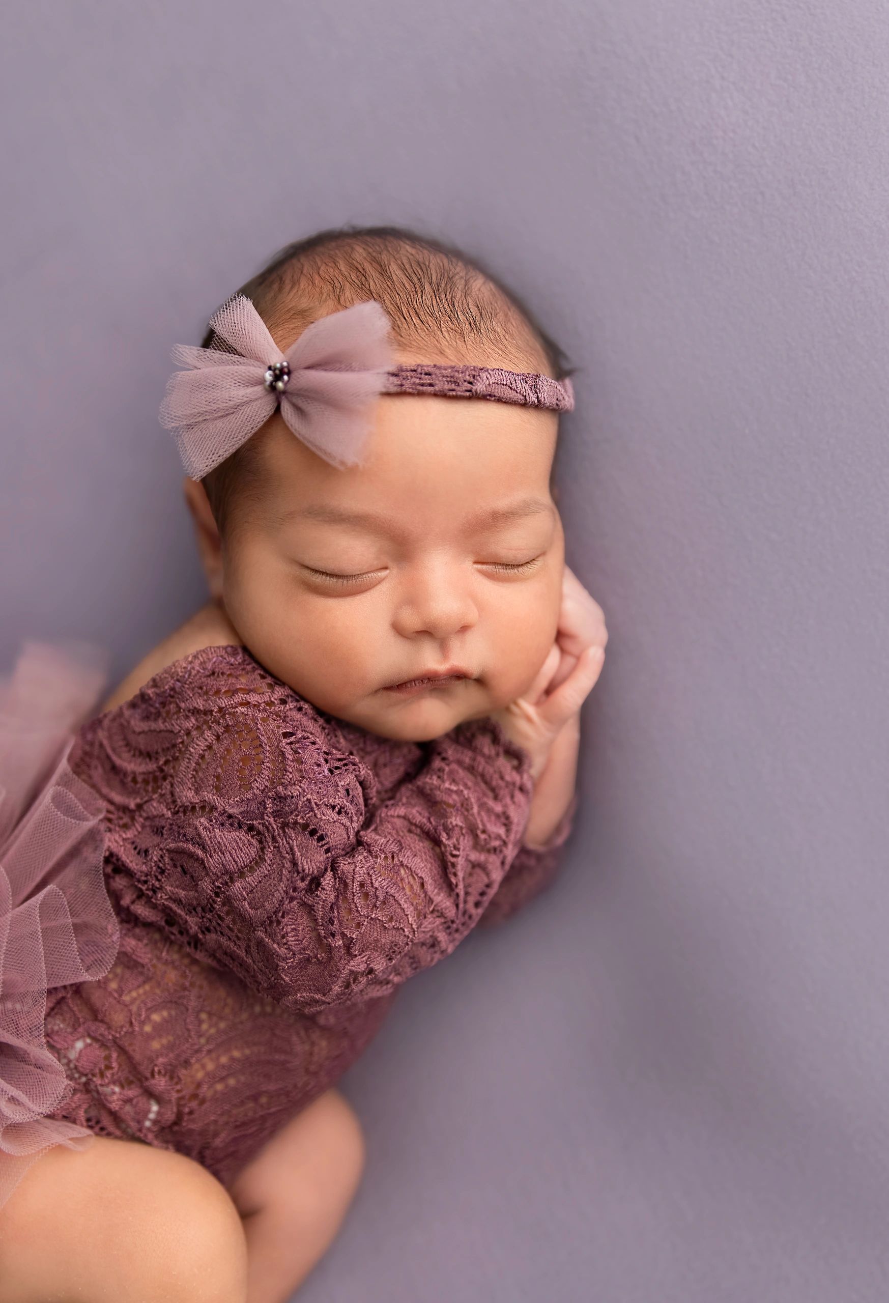 Sleeping newborn baby in purple lace outfit with matching headband and bow.