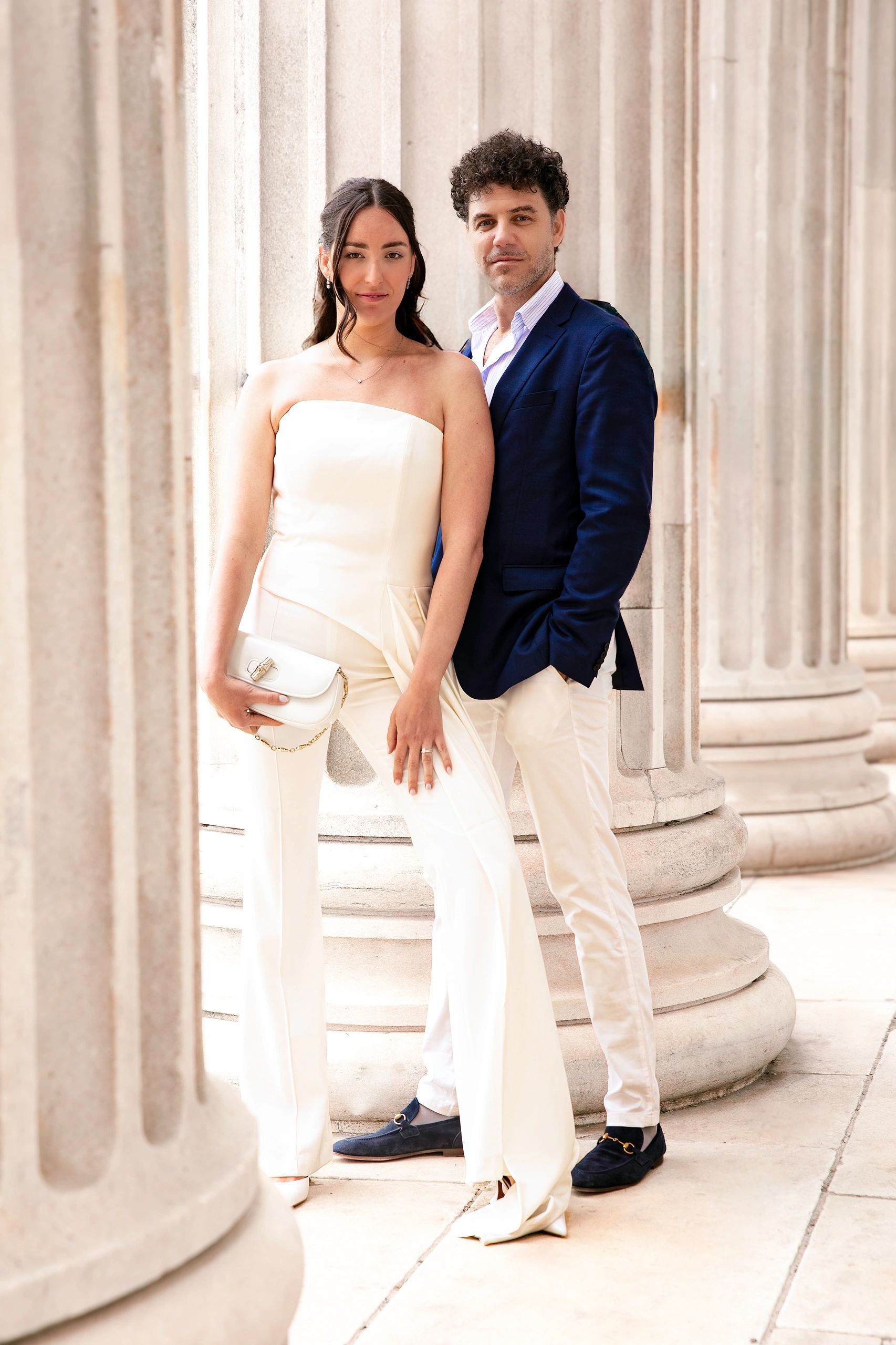 Elegant couple posing between classical stone columns, dressed in chic white and navy attire.