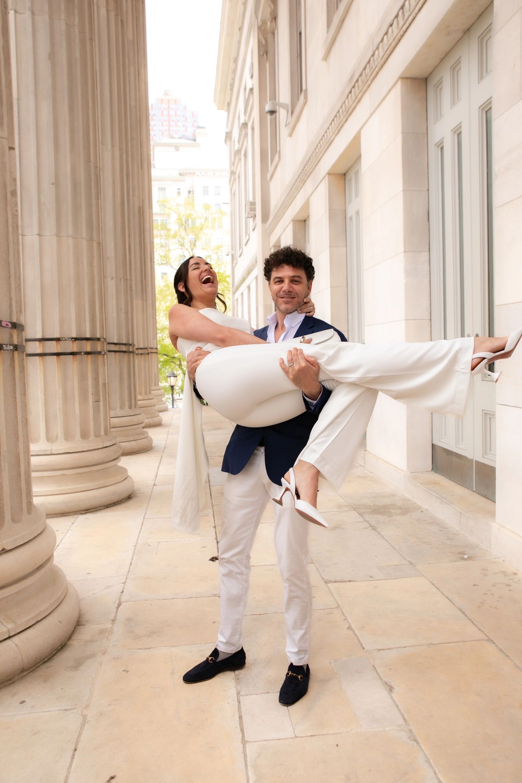Man in a suit carrying a laughing woman in white pants and heels outdoors.