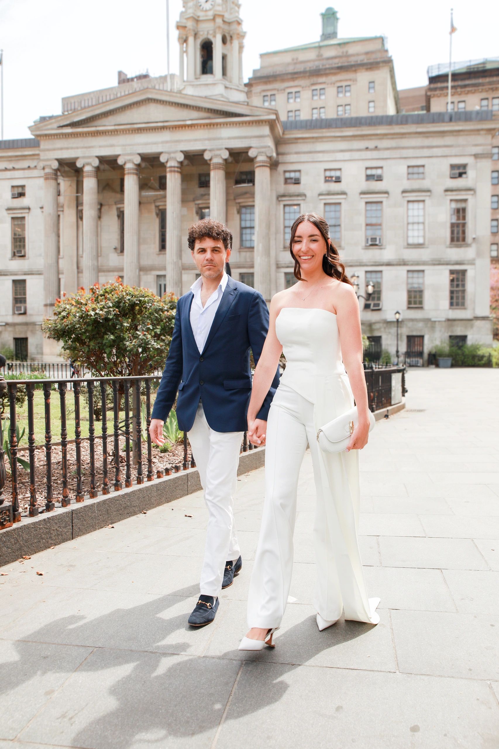 Couple walking hand in hand outside a historic building, dressed elegantly.
