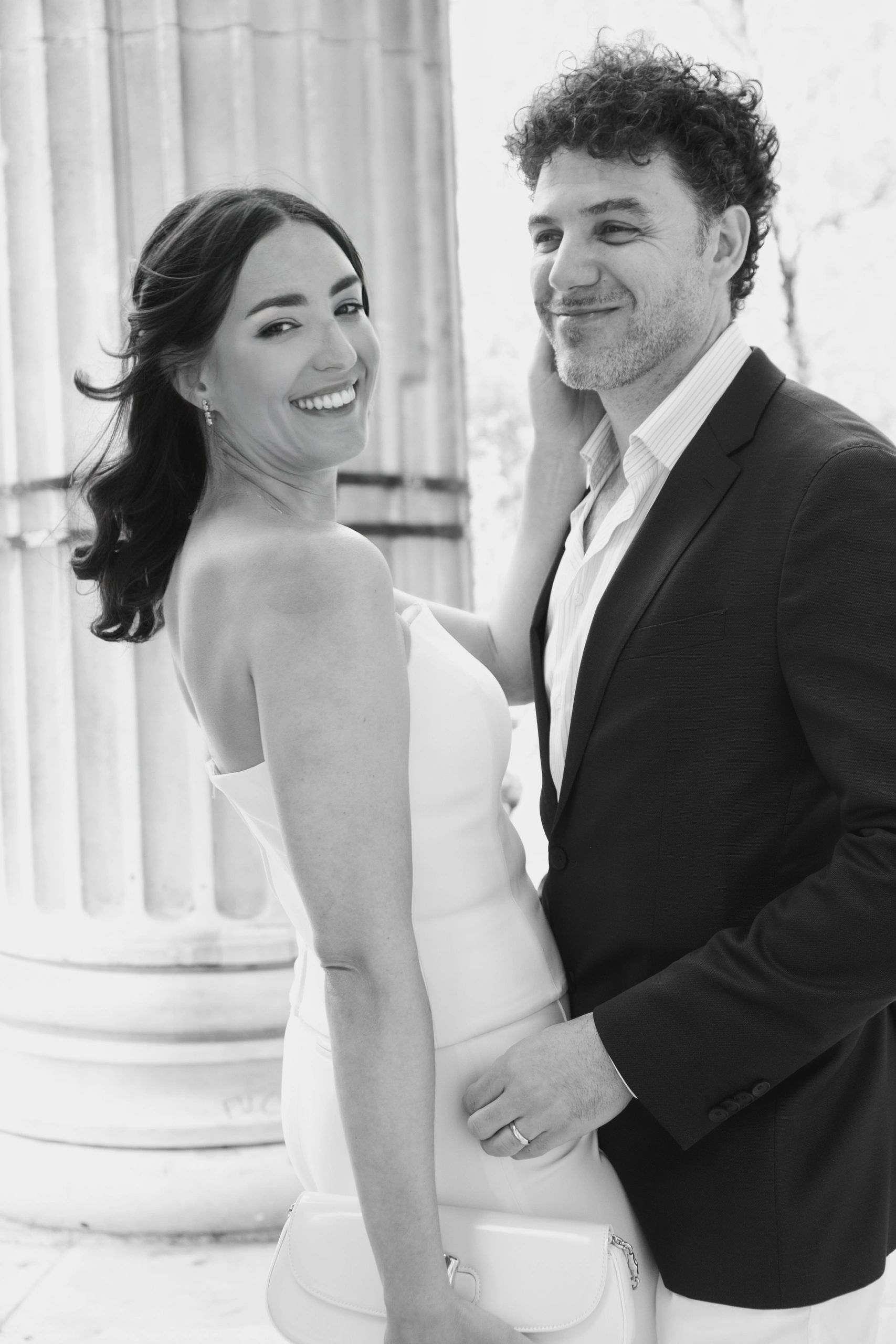 Elegant couple smiling beside classical columns in black and white.