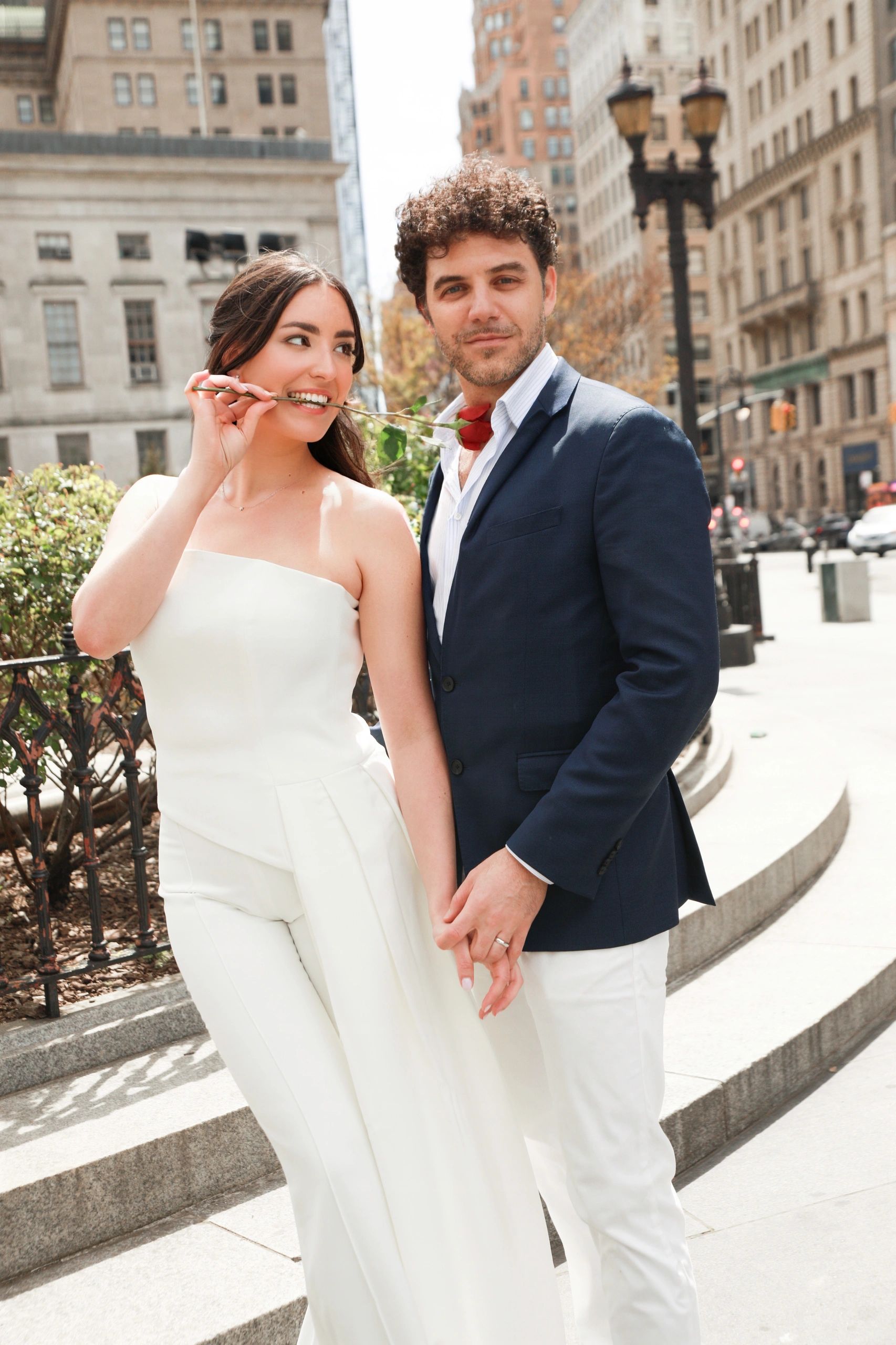 Couple holding hands with roses in a city setting, dressed stylishly.
