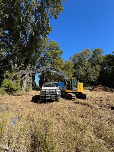 A yellow excavator and white truck in a clearing surrounded by trees under a blue sky.