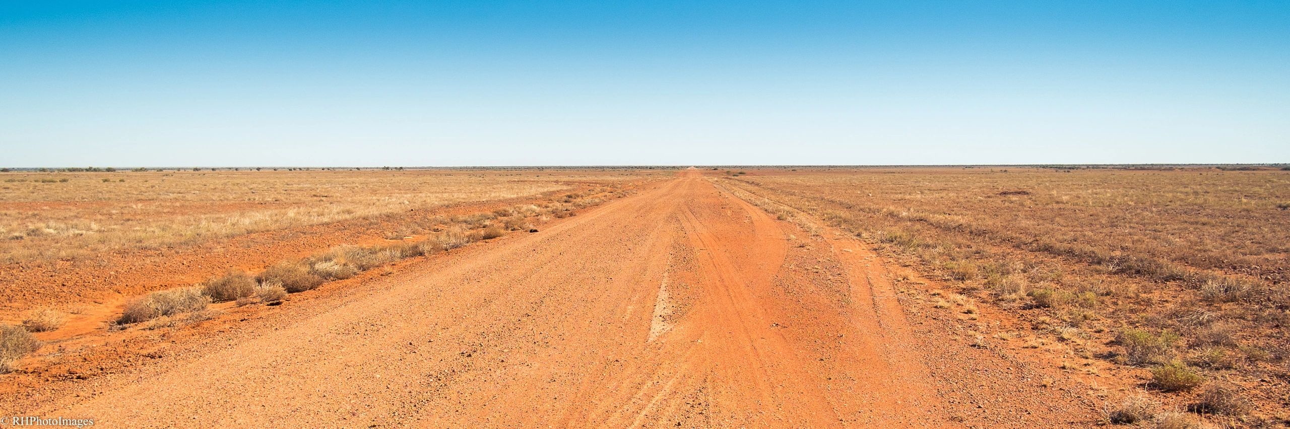 A long dirt road stretches through a dry, flat landscape under a clear blue sky.