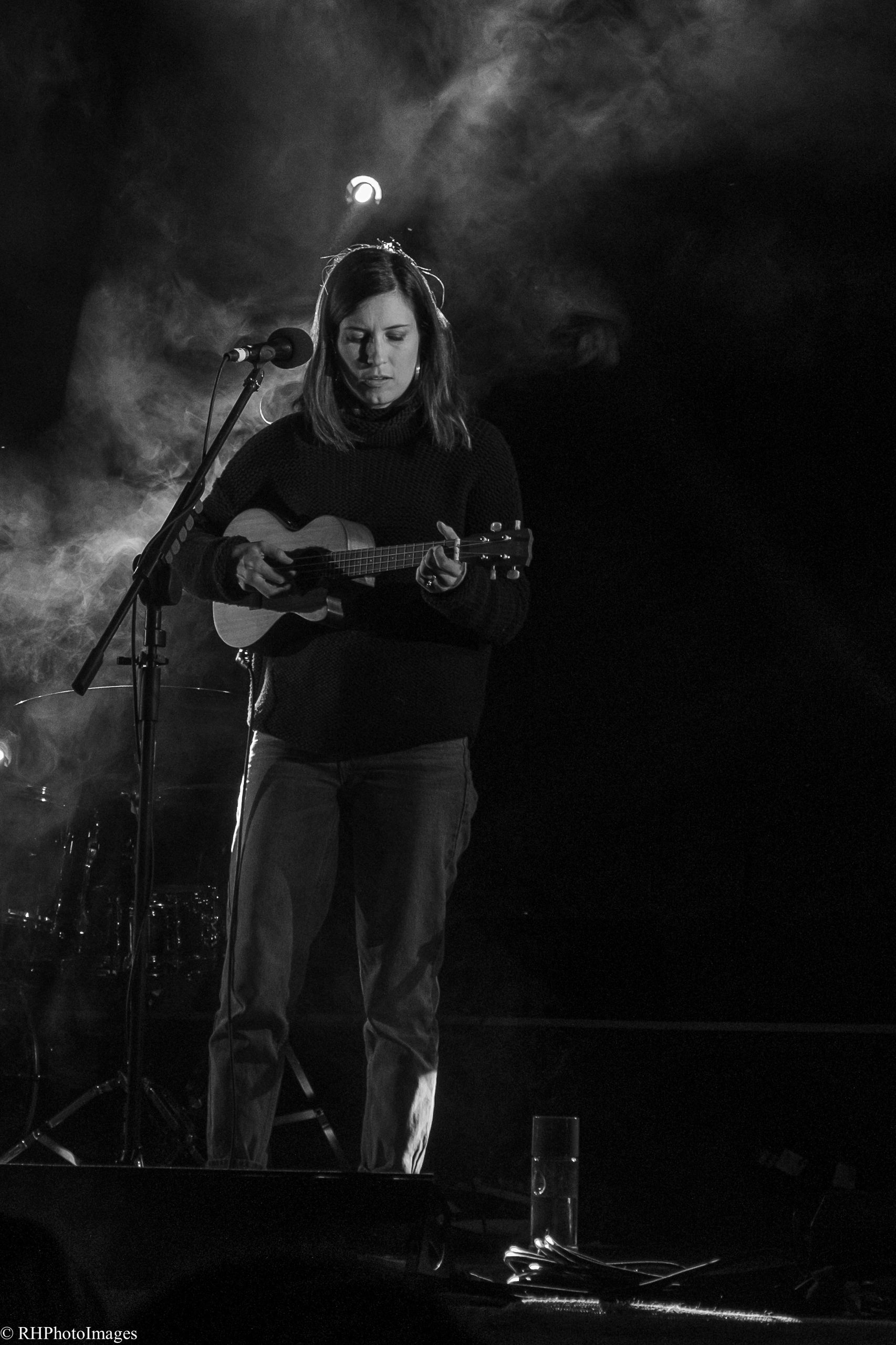 Woman playing ukulele on stage with moody lighting and smoke.