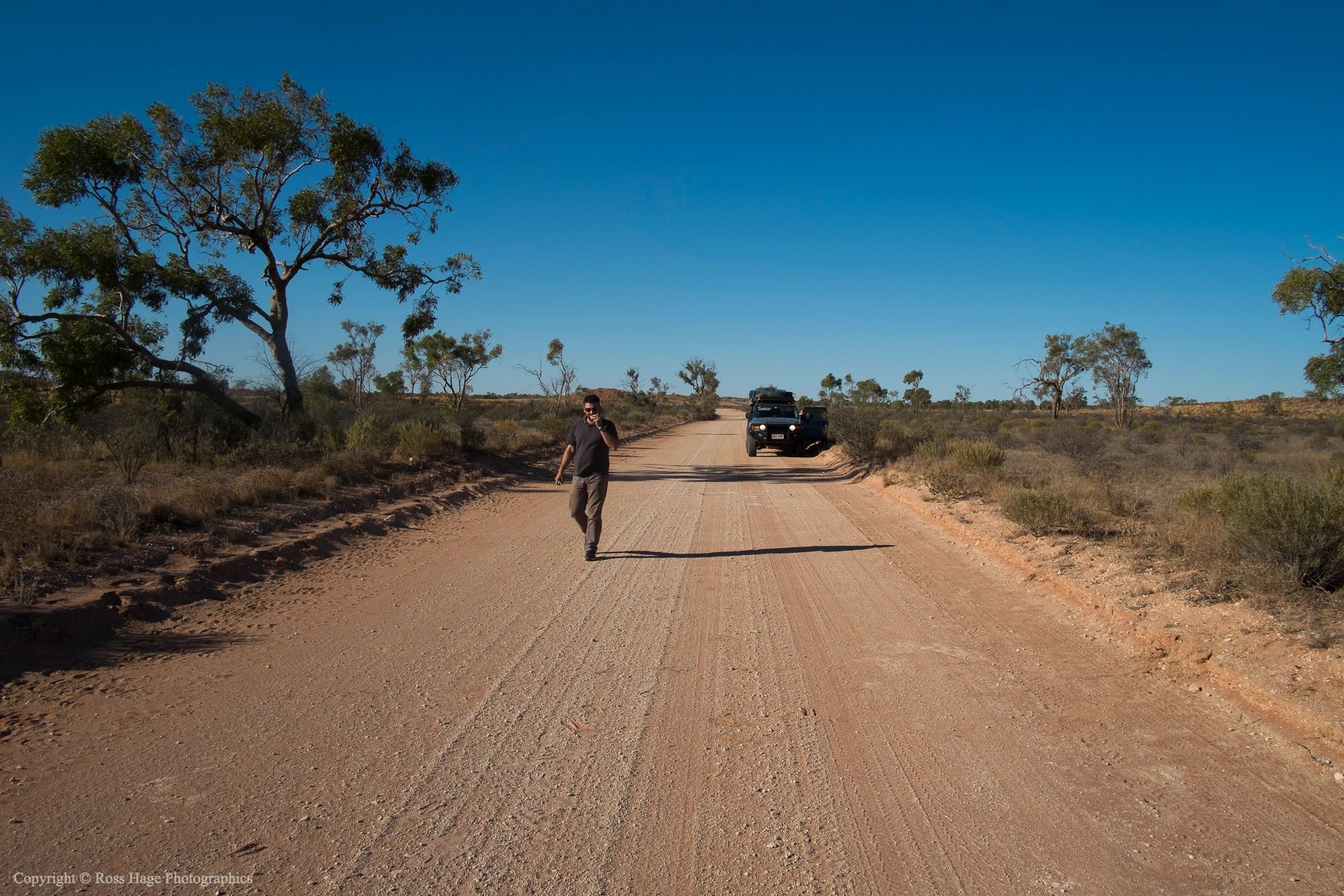 A man walks on a dirt road near a parked SUV in a dry, open landscape.