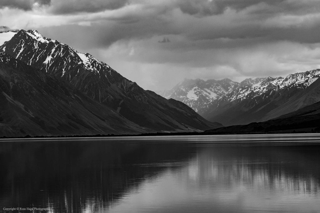 Black and white photo of snow-capped mountains reflected in a calm lake under cloudy skies.