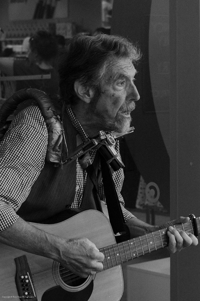 An elderly man passionately playing guitar and harmonica in black and white.
