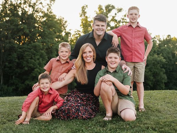 A husband, wife, and four sons smiling together in a park.