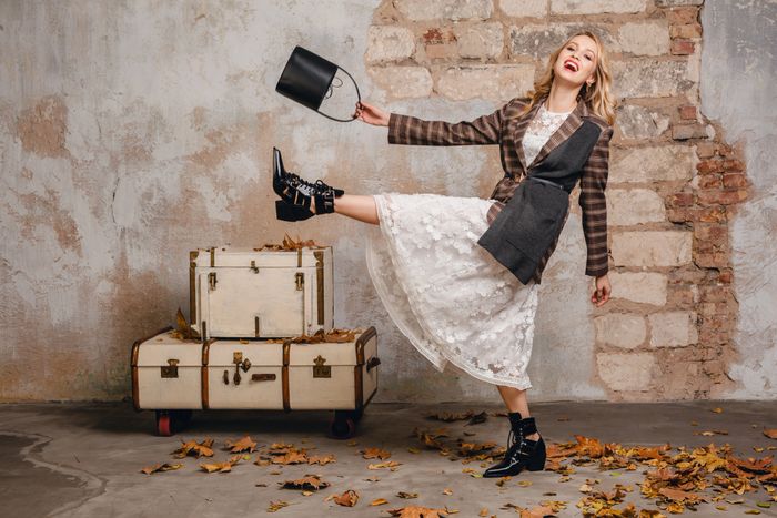 A joyful woman in a stylish outfit poses playfully on one leg with a handbag.