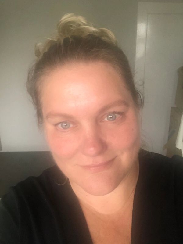 A close-up selfie of a woman with light eyes and a soft smile in a softly lit room.