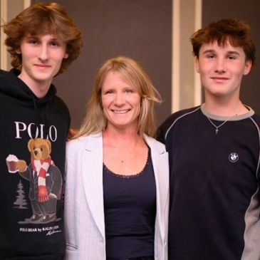 A smiling woman stands between two teenage boys in casual clothing indoors.