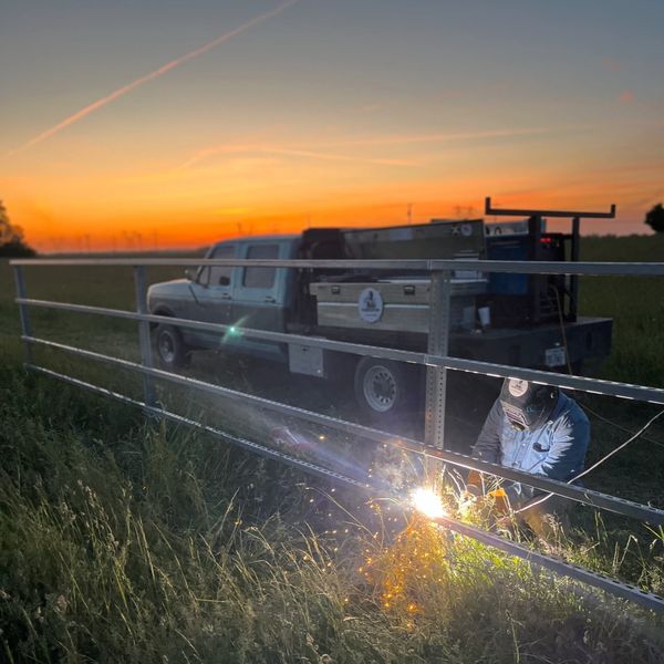 Person welding a metal fence at sunset with a pickup truck nearby.