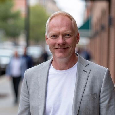 Smiling man in a light gray blazer and white shirt on a city sidewalk.