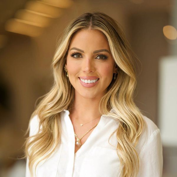 Smiling woman with long blonde hair wearing a white blouse indoors.