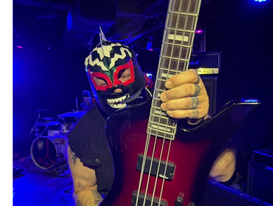 Musician in a colorful lucha libre mask playing a bass guitar on stage.