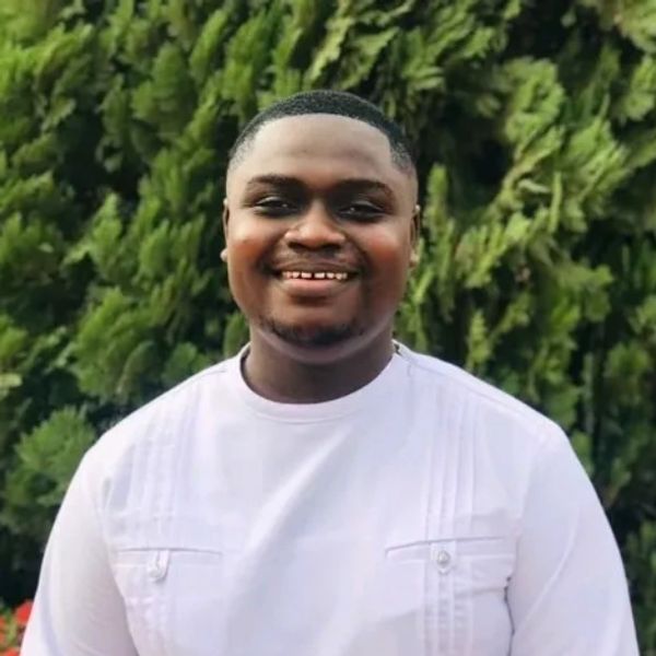 A smiling man in a white shirt stands outdoors with green foliage behind him.