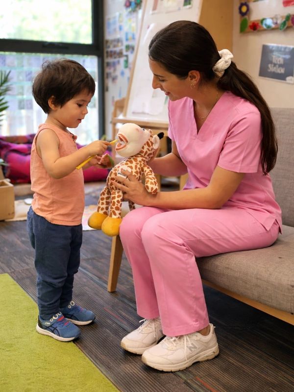 A child practices brushing a giraffe toy's teeth with a caregiver's help.